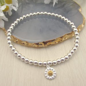 May include: Silver beaded bracelet with a daisy charm. The bracelet features small, round silver beads strung together. The daisy charm has a silver outer layer and a gold center.