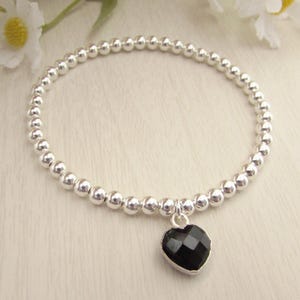 May include: Silver beaded bracelet featuring a black heart-shaped charm. The charm has a faceted surface, and the bracelet is made of small, round silver beads. The bracelet is displayed on a light-coloured surface.