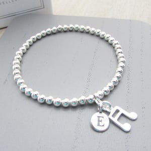 Könnte beinhalten: Silbernes Perlenarmband mit einem Notenschlüssel-Anhänger und einem runden Anhänger mit dem Buchstaben "E". Das Armband liegt auf einer grauen Oberfläche. Der Schmuck ist wahrscheinlich aus Sterlingsilber.