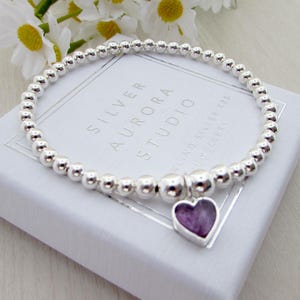 Puede incluir: Una pulsera de cuentas plateadas con un dije en forma de corazón. El dije tiene una piedra morada engastada en un marco plateado. La pulsera descansa sobre una caja blanca con el texto "SILVER AURORA STUDIO".