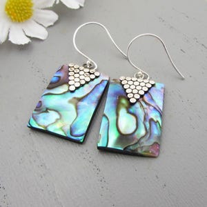 Può includere: Un paio di orecchini rettangolari con conchiglie di abalone iridescenti nei toni del blu, verde e viola. Gli orecchini hanno ganci in metallo color argento e un design triangolare nella parte superiore. Gli orecchini sono esposti su una superficie grigia.