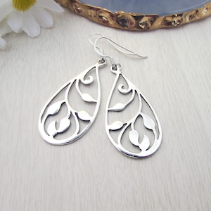 May include: Silver teardrop earrings featuring a cut-out leaf pattern. The earrings have a hook fastening and are shown on a light wooden surface. These earrings are a delicate and elegant accessory, perfect for everyday wear.