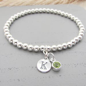 May include: A silver beaded bracelet with a charm featuring the letter K and a small green gemstone.