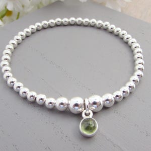 May include: Silver beaded bracelet with a small, round, green gemstone charm. The bracelet is made of multiple small, round silver beads. The charm is a light green colour and hangs from a silver loop.
