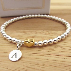 Könnte beinhalten: Ein silbernes Armband mit Perlen, einem goldenen Herz-Anhänger und einem silbernen Anhänger mit dem eingravierten Buchstaben A.