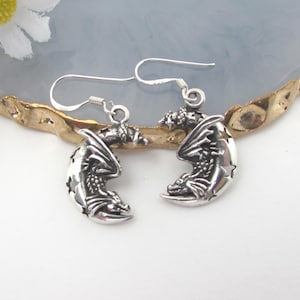 May include: A pair of silver-toned dangle earrings featuring a crescent moon design with a dragon perched on the curve. The moon shape includes cut-out star details and the earrings are attached to standard silver-toned ear wires for hanging.