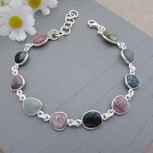 May include: A silver bracelet with a series of irregularly shaped, colourful gemstones. The stones are set in individual silver bezels and linked with small silver chain segments. The gemstones are various shades of pink, green, and blue.