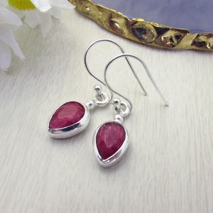 May include: Silver drop earrings featuring teardrop-shaped, deep red gemstones. The earrings have a simple, elegant design with a polished finish. The gemstones are set in a silver bezel.