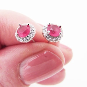 Könnte beinhalten: Ein Paar silberne Ohrringe mit einem runden, roten Rubin-Edelstein, der von einem Heiligenschein aus kleineren, klaren Edelsteinen umgeben ist.