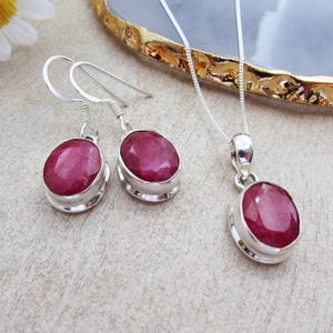 Peut inclure: Un ensemble de bijoux comprenant une paire de boucles d'oreilles et un collier pendentif assorti. Chaque pièce présente une grosse pierre précieuse rouge foncé de forme ovale, sertie dans un sertissage en argent. Les boucles d'oreilles sont à crochet, et le collier a une chaîne fine.