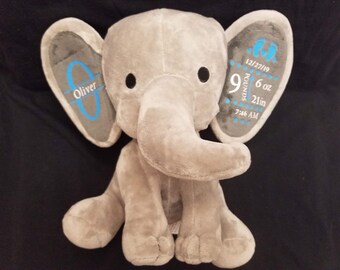 Op de afbeelding: Grijze pluche olifant knuffel met blauwe geborduurde details. Het ene oor zegt "Oliver" en het andere oor zegt "9 pond 6 oz, 21 inch, 7:46 AM, 12/27/19".