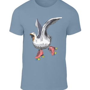 Puede incluir: Una camiseta azul claro con un gráfico de una gaviota con patines de ruedas rojos. La gaviota está volando con las alas extendidas y la boca abierta en un grito.