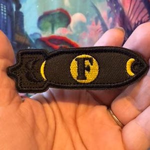 May include: Black embroidered patch in the shape of a bomb. The patch features a yellow letter "F" in a circle, with yellow crescent moon shapes on each end. The patch is held in a hand.
