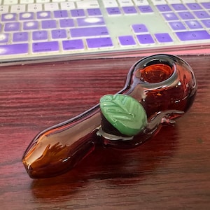 May include: A brown glass pipe with a green leaf design. The pipe has a bowl on one end and a mouthpiece on the other.