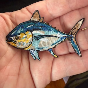 May include: Embroidered patch of a tuna fish. The patch features a detailed design with shades of blue, yellow, and brown. The fish has a streamlined body, fins, and a tail. The patch is likely for clothing or accessories.