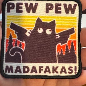 May include: Square patch with a black cat silhouette holding two guns, with the words "PEW PEW MADAF*KAS!" in white. The background features a sunset design with orange, yellow, and red hues, and a forest silhouette.