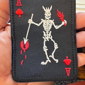 May include: Black Ace of Spades patch featuring a white skeleton holding a spear and a red flame. The patch has red spade symbols and the letter "A" at the top and bottom corners.