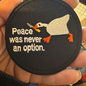 May include: A round, black embroidered patch featuring a white goose with an orange beak and feet, holding a knife. The text "Peace was never an option" is embroidered in white.
