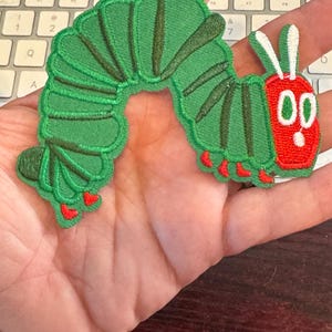 May include: A green caterpillar patch with a red head and feet. The caterpillar has a segmented body with dark green stripes and red accents. The patch is likely for clothing or accessories.