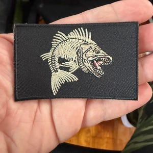 May include: Black rectangular patch featuring an embroidered fish skeleton with an open mouth and visible teeth. The fish is in a light beige color, contrasting against the black background. The patch is likely for clothing or accessories.