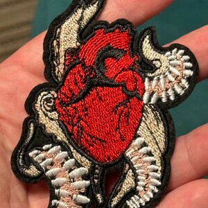May include: Embroidered patch featuring a red heart with black outlines and detailed stitching. The heart is surrounded by stylized wings in shades of beige and white, all set against a black background. This patch is suitable for jackets, bags, or other fabric items.
