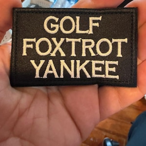 May include: Black rectangular patch with white embroidered text that reads "GOLF FOXTROT YANKEE". The patch is held in a person's hand, with a wooden surface in the background.