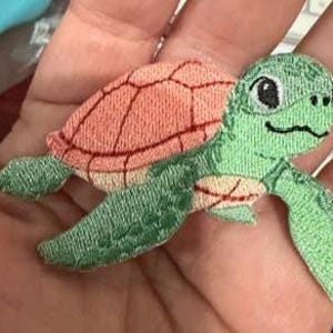 May include: Embroidered sea turtle patch featuring a green head and flippers, and a coral-colored shell. The patch is detailed with black eyes and stitching, and is likely for clothing or accessories.
