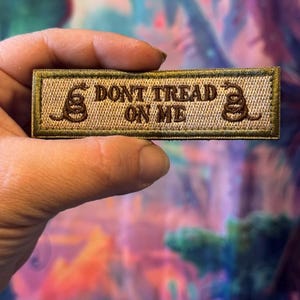 May include: A rectangular patch with the text "DON'T TREAD ON ME" embroidered in brown thread. The patch features a coiled snake on each side of the text. The border is olive green, and the background is tan.