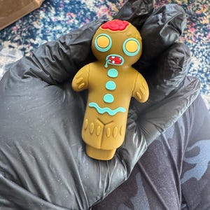 Puede incluir: Un juguete de hombre de jengibre marrón con una cabeza de glaseado rojo y blanco, ojos amarillos y una sonrisa de glaseado azul. El juguete tiene un diseño de glaseado azul en el cuerpo y una fila de puntos marrones en la parte inferior.