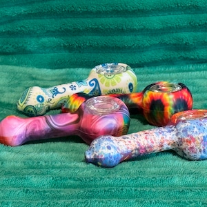 May include: Four colorful silicone smoking pipes with glass bowls. The pipes have different designs, including paisley, tie-dye, and marble.