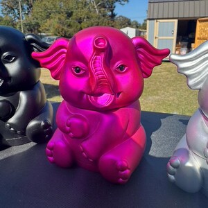 May include: A vibrant pink elephant figurine with a metallic sheen, smiling with its trunk raised. The elephant is one of three, with a black and a silver elephant also visible. The figurines are sitting on a dark surface.