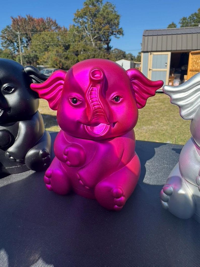 May include: A vibrant pink elephant figurine with a metallic sheen, smiling with its trunk raised. The elephant is one of three, with a black and a silver elephant also visible. The figurines are sitting on a dark surface.
