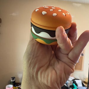 May include: A novelty burger-shaped container with a bright orange top dotted with white sesame seed details. The burger has layers of brown, white, and green, resembling a classic hamburger. The container is held in a hand.