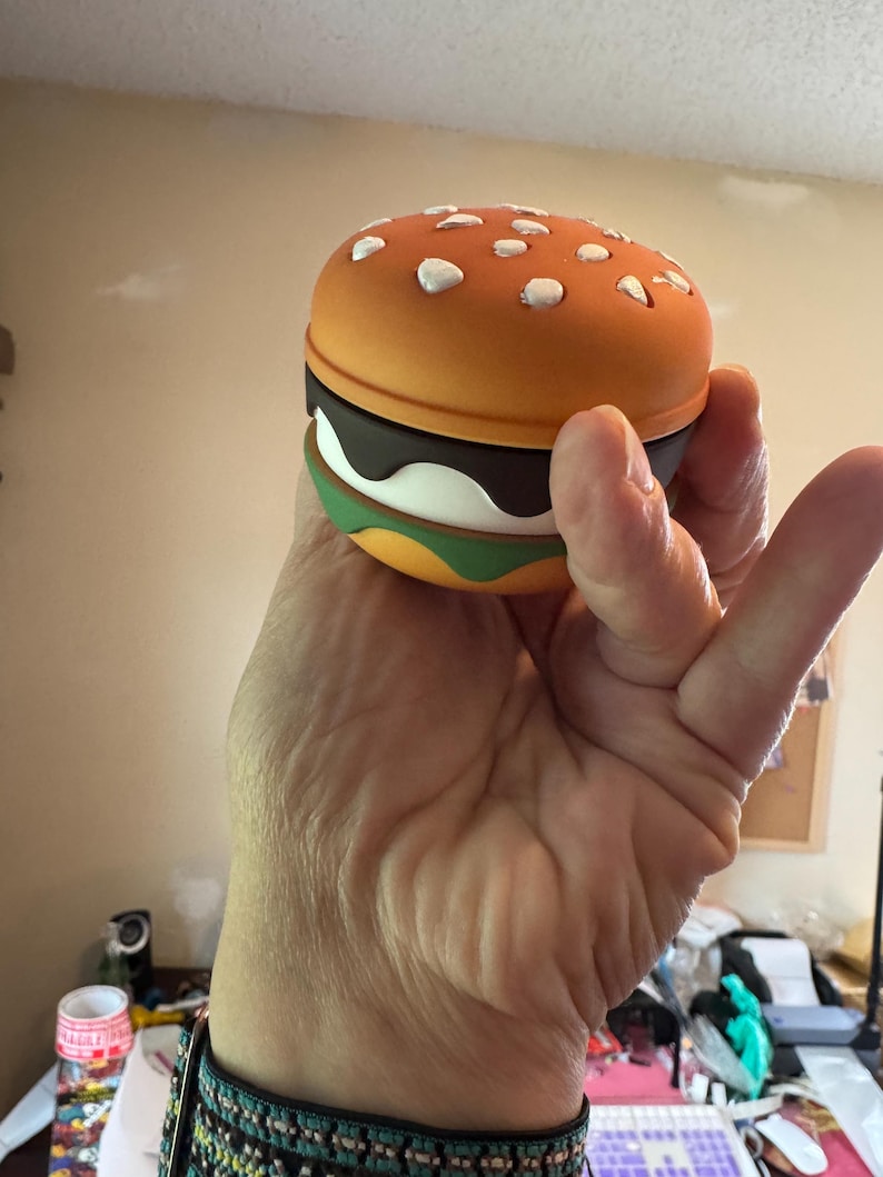May include: A novelty burger-shaped container with a bright orange top dotted with white sesame seed details. The burger has layers of brown, white, and green, resembling a classic hamburger. The container is held in a hand.