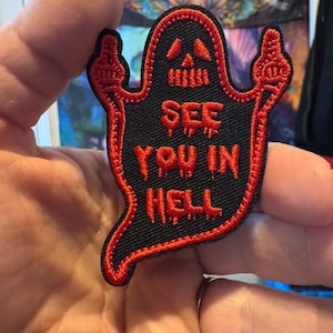 May include: Black and red embroidered patch in the shape of a ghost. The ghost has a skull-like face and is giving the middle finger with both hands. The text "SEE YOU IN HELL" is embroidered in red.