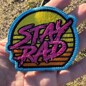 May include: Embroidered patch with the words "STAY RAD" in pink, outlined in black, set against a yellow and orange sunset design. The patch has a blue border and is likely for clothing or accessories.