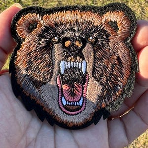 May include: Embroidered bear patch with a snarling expression. The patch features brown, black, and white thread detailing the bear's fur, teeth, and open mouth. The patch has a black border and is held in a hand.