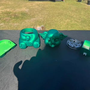 May include: A collection of green, novelty items, including an elephant-shaped container, a lid, a grinder, and a small box. The elephant has wings and a trunk. The items are arranged on a dark surface.