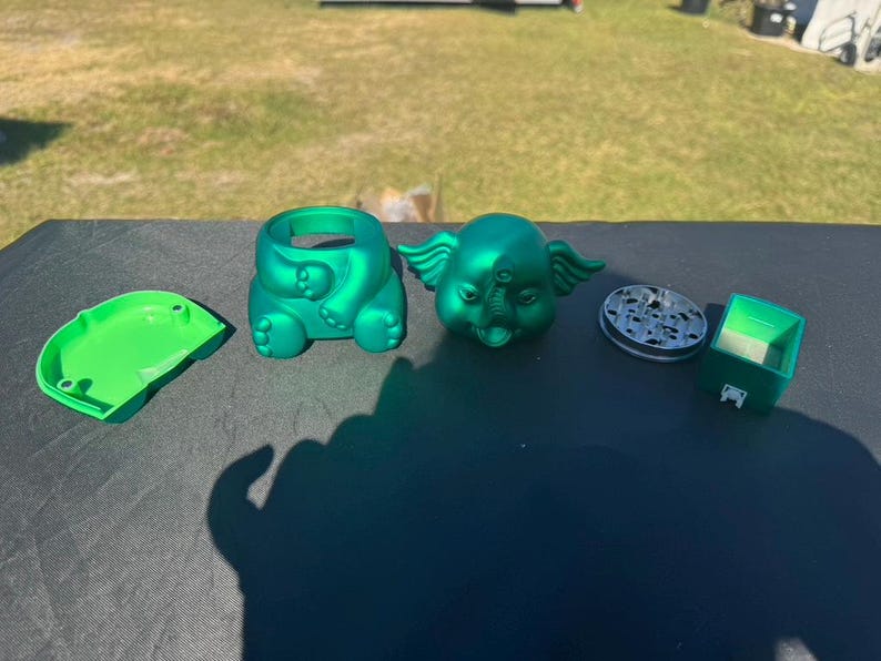 May include: A collection of green, novelty items, including an elephant-shaped container, a lid, a grinder, and a small box. The elephant has wings and a trunk. The items are arranged on a dark surface.