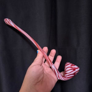 Puede incluir: Una pipa de vidrio con un diseño a rayas rojas y blancas. La pipa tiene un tallo curvo y un cuenco redondeado. Los colores están mezclados, creando un patrón único. La pipa está sobre un fondo negro.