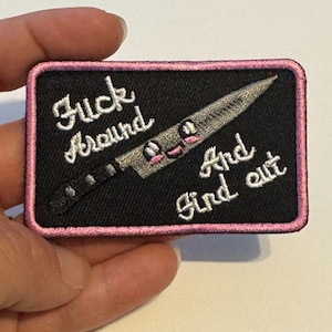 May include: Embroidered patch with a black background and a pink border. The patch features a cartoon knife with a face and the words "Fuck Around And Find Out" in white script.