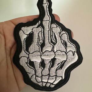 May include: A black and white embroidered patch of a skeleton hand giving the middle finger.