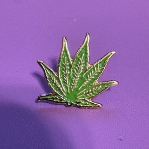 May include: A gold-colored pin with a green marijuana leaf design.
