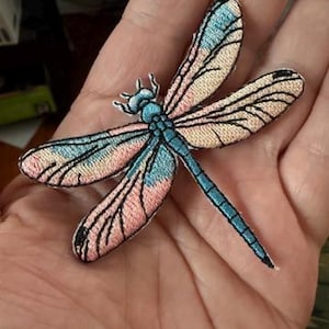 May include: Embroidered dragonfly patch featuring a detailed design with blue, pink, and black accents. The dragonfly has delicate wings and a slender body. This patch is suitable for clothing, bags, or other fabric items.