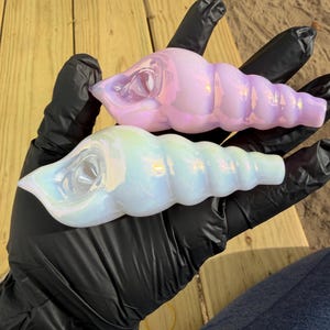 May include: Two iridescent glass seashell sculptures, one pink and one white, are held in black gloved hands. The shells have a pearlescent finish and a ridged texture, resembling natural sea shells.