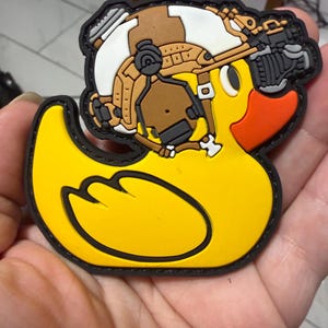 May include: A yellow rubber duck patch with a black outline. The duck has an orange beak and is wearing a brown, white, and gray tactical helmet with a gun attached. The patch is held in a person's hand.