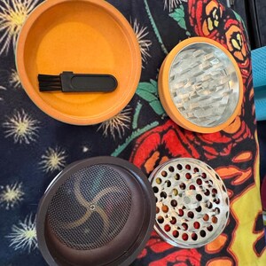 May include: An orange and silver herb grinder with multiple components. The grinder is disassembled, revealing the different parts, including a brush, a mesh screen, and a grinding chamber. The background is a colorful floral pattern.