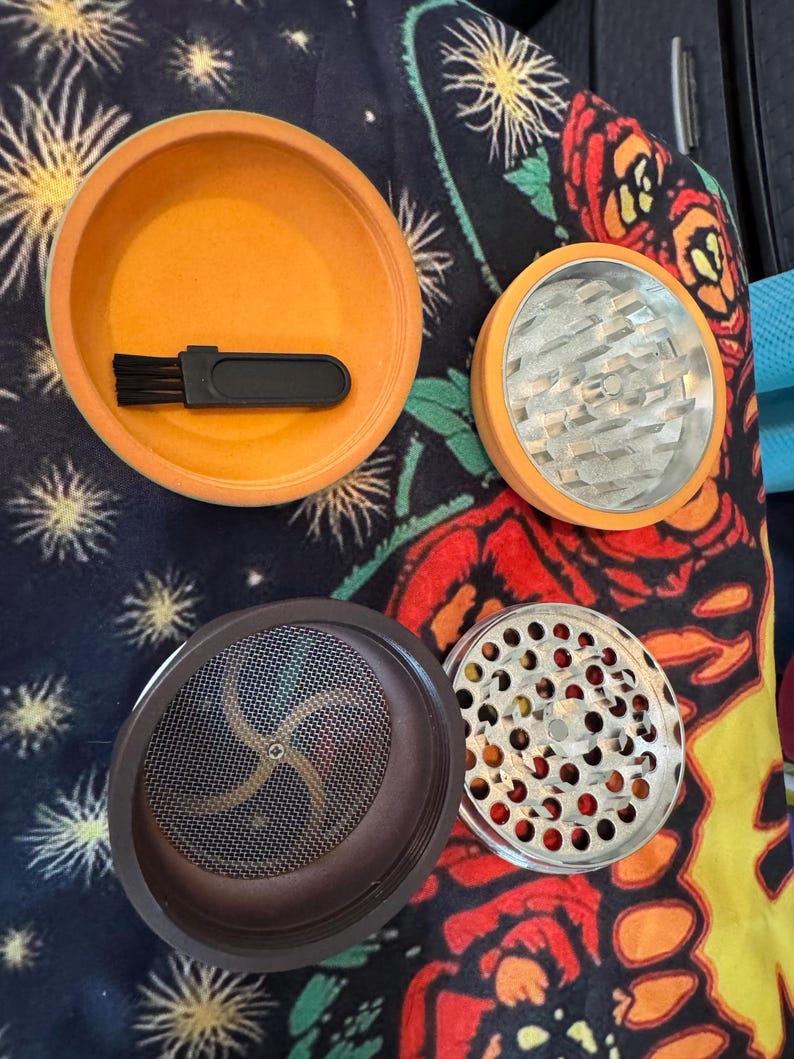 May include: An orange and silver herb grinder with multiple components. The grinder is disassembled, revealing the different parts, including a brush, a mesh screen, and a grinding chamber. The background is a colorful floral pattern.