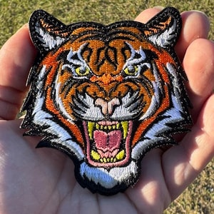 May include: Embroidered tiger head patch with orange, black, white, and yellow details. The tiger is snarling, with its mouth open, revealing its teeth and pink tongue. The patch is likely for clothing or accessories.