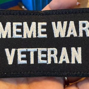 May include: Black rectangular patch with white embroidered text reading "MEME WAR VETERAN". The patch is likely made of fabric and has a stitched border. The text is in a bold, sans-serif font.